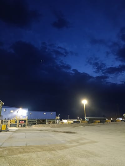 This image depicts an industrial outdoor scene at dusk or night in Қопа ауылдық округі, Kazakhstan. The sky is a deep, dark blue with visible clouds, suggesting twilight or night.
The primary subjects are several large, blue modular buildings or shipping containers arranged in a line. They are illuminated by artificial lights, with one building prominently featuring a bright light source on its exterior. A yellow metal staircase with railings leads up to one of the buildings, suggesting access for personnel. To the left of this building, a yellow piece of equipment, possibly a forklift or construction machinery, is visible.
In the middle ground, a tall lamppost with a bright, glowing light illuminates the paved and gravelly ground. Several yellow and black striped barriers and stacks of tires are placed along the edge of the paved area, possibly for traffic management or safety. Further in the background, more structures and lights can be seen, indicating a larger industrial complex or work site. The overall setting appears to be a functional, possibly construction or industrial yard, with a sense of activity even in the low light conditions. There are no people clearly visible in the scene. The mood is functional and industrial, with the artificial lights creating a stark contrast against the darkening sky.