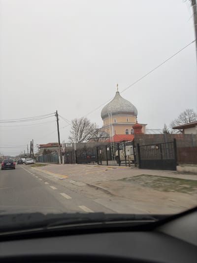 The image presents a street-level view captured from inside a vehicle on an overcast day in 2 Mai, Romania. The primary subject is the Biserica Ortodoxă "Sfinții Arhangheli Mihail și Gavriil," characterized by its prominent grey, onion-shaped dome topped with a golden cross. The church structure itself is light beige with visible arched windows and a section featuring a red-tiled roof, with a smaller red-domed structure partially visible behind a gate.

The foreground displays the dashboard and lower windshield area of the capturing vehicle. The street consists of paved asphalt with white dashed lane markings. To the immediate right of the street is a paved sidewalk or parking area, marked with yellow lines indicating individual parking spaces, separated from the road by concrete curbing.

A tall black metal fence, featuring decorative elements and a large gate, encloses the property adjacent to or containing the church. A white sign with black lettering, partially legible, displays "CAZARE" (Accommodation) and a phone number. Behind this gate, a white utility van and another dark-colored vehicle are visible. Utility poles with multiple overhead power lines are positioned along the street.

On the left side of the street, several vehicles are present, including a dark-colored station wagon or SUV and a lighter-colored sedan, indicating traffic or parked cars. Further down the street, residential or commercial buildings with red-tiled roofs are visible amidst bare deciduous trees, suggesting late autumn, winter, or early spring. No individuals are visible in the scene.