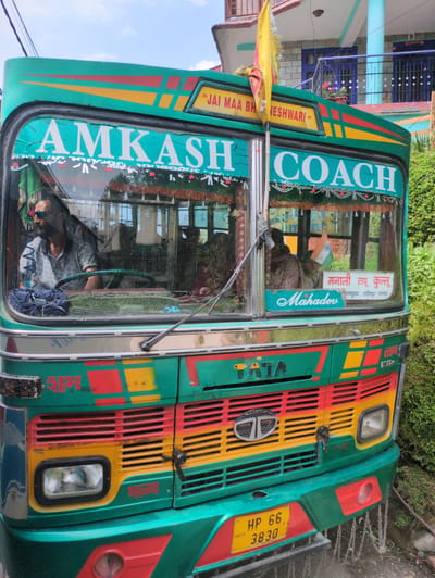 This media file is a photograph of a vibrantly colored bus parked outdoors in a setting that suggests a rural or mountainous area, likely in India given the text and license plate. The bus is painted primarily in green with striking accents of red and yellow, featuring bold lettering on its exterior.
The subjects in the image include the bus itself, which is the central object, and a person, likely the driver, visible through the windshield. The driver is wearing sunglasses and appears to be looking to their left. The scene is set against a backdrop of lush green foliage, suggesting it might be daytime with good weather. Behind the bus, glimpses of a building with blue and white elements and a tiled roof are visible.
Notable details include the text displayed on the bus. On the side, "AMKASH COACH" is written in large white letters on a teal background, with "JAI MAA BH JESHWARI" in a yellow rectangle above it. The front of the bus has a colorful grille with "TATA" branding. The windshield also displays text in different languages, including "मनाली टापू कुल्लू" (Manali Top Kullu) and "Mahader". A license plate reading "HP 66 3830" is also visible. A small Indian flag is displayed inside the bus's cabin.
The overall impression is that of a well-decorated public transport vehicle, possibly operating on a route connecting to tourist destinations like Manali. The bus's presence in this outdoor, natural setting hints at its role in transportation within a region.