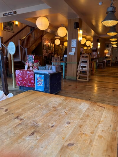 This indoor shot showcases a warm ambiance with wooden floors and a visible staircase. A wooden table dominates the foreground, leading to an obscured view of the interior. A display features Valentine's-themed items, including a red gift-wrapped box and decorative reindeer. Various light fixtures illuminate the space, with spherical lamps on the left and industrial-style lamps hanging from the ceiling. Upward signage indicates restroom directions. The blurred depth of field suggests a cozy, slightly cluttered environment.