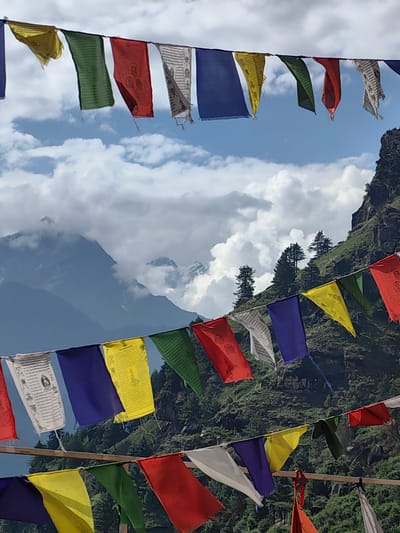 The image shows prayer flags strung across a mountainous landscape in Siumsa, India. The flags are colorful and feature various patterns and symbols. They are attached to a wire or rope and hang loosely in the wind. In the background, there are towering mountains with patches of snow and lush green vegetation. The sky is partly cloudy, with dramatic cloud formations adding to the scenic beauty of the location. The image captures a serene and spiritual atmosphere, characteristic of the Himalayas.