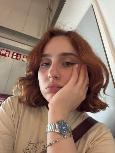 A young woman with auburn, curly hair rests her face in her hand, appearing contemplative. She wears a beige t-shirt and a silver watch with a dark blue face. A brown shoulder strap is visible. In the background, a wall with signs depicting fire safety equipment is visible. The location is a passenger train in Lautzenhausen, Germany.