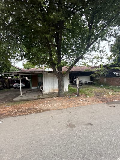 The image depicts a modest, single-story structure in Ajuen, Indonesia, with a corrugated metal roof. The building appears to be a residence or small business, constructed with white wooden walls. To the left, a carport shelters a silver car. A motorcycle is parked in front of the building, near a small table and chairs. The surrounding area is overgrown with grass and scattered with debris, including plastic bottles. A large tree with dense foliage dominates the foreground, partially obscuring the structure. A paved road runs in front of the property, with fallen leaves and dirt on the roadside. The sky is overcast, suggesting a cloudy day. There are no people visible in the image.