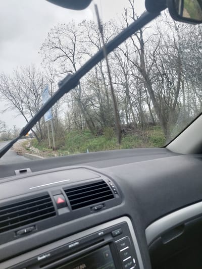 The image shows a view from inside a car, looking out the windshield. A windshield wiper is visible. Outside the car, there are bare trees and some greenery, suggesting a rural or park-like environment. A blue signpost is also visible. Based on the environment with trees and nature, and signpost, it could be on the outskirts of the town or on a road leading into or out of Gorski Izvor.