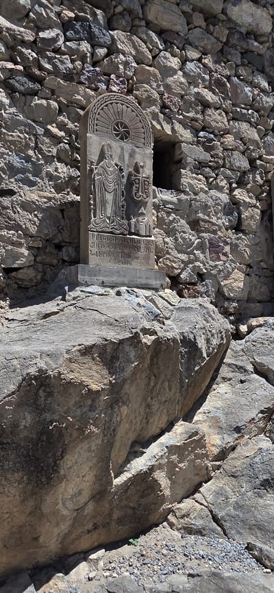 The image depicts a stone monument with bas-relief carvings, positioned in front of a rough stone wall. The monument is tall and rectangular, topped with a decorative arched section featuring a sun-like motif. Below this, a figure in religious attire, possibly a bishop, is carved, standing to the left of a large cross with an architectural element at its base. Inscriptions are visible in Armenian script below the figures. The monument rests on a concrete base, which is situated on a large, weathered rock. The background is composed of a wall constructed from irregular stones of varying sizes and colors. A dark rectangular opening is visible in the stone wall to the right of the monument. The ground around the base of the monument is rocky and uneven. The overall environment appears to be outdoors, with sunlight casting shadows on the stone structures. This monument is identified as being in Lusarat, Armenia.