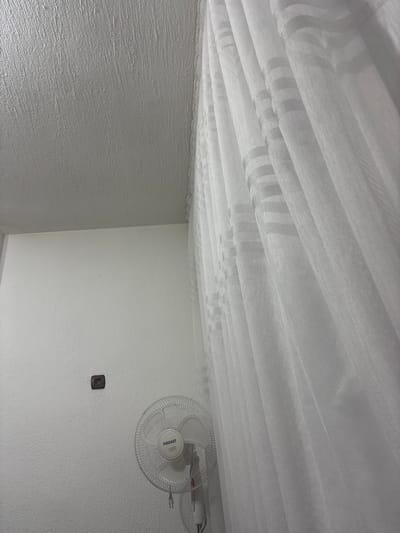 The image shows an interior view of a room. The ceiling is covered with white fabric, possibly mosquito netting or curtains. The walls are white with a slightly textured surface. A white electric fan is mounted on the right wall, above a small brown electrical outlet. The image orientation suggests that the photographer is looking upwards. There is no clear indication of the location in Grohotno, Bulgaria, or any specific details about people or actions within the scene.