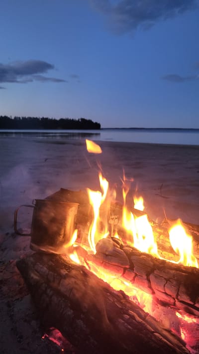 A campfire burns on a sandy beach near the body of water in Zaostrov'e, Russia. The sky is a deep blue, indicating twilight. The fire is comprised of logs, with visible flames and glowing embers. To the left of the fire, a metal mug with a handle is positioned on the sand. The background shows a calm body of water with a treeline visible in the distance. The scene is quiet and serene.