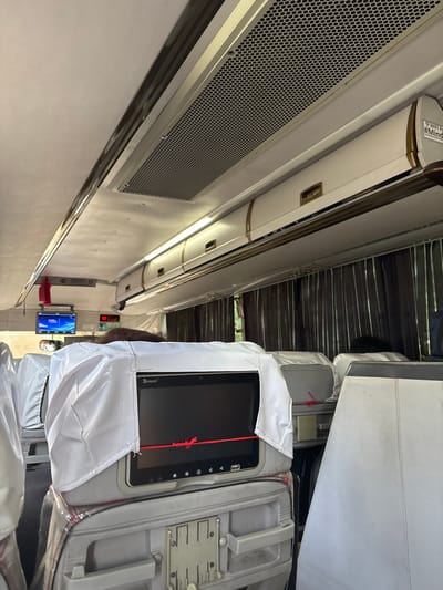 This is an interior view of a bus in Santa Cruz, Philippines, looking forward. The ceiling is cream-colored, with a large air conditioning vent running horizontally along the top. Below the vent are overhead storage compartments with gold accents. The seats in the foreground are covered in white plastic sheeting. A screen, labeled "buspad," is visible on one of the seats, displaying a red line across a black background. To the left, a small blue screen displaying text is mounted on the wall, and further down, a red digital display shows "00." Dark brown curtains hang along the side of the bus. A person's dark hair is visible above the seats.