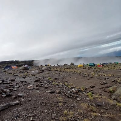 The image depicts a high-altitude mountain camp at Gilman's Point, Tanzania, under an overcast and misty sky. The terrain is barren, characterized by dark brown soil, scattered grey and black volcanic rocks, and sparse patches of low-lying, dull green vegetation. A prominent horizontal band of white mist or low clouds obscures the distant horizon, while heavy grey, streaked clouds dominate the sky above. Approximately two dozen multi-colored camping tents (including blue, orange, red, yellow, green, and grey) are distributed across the rocky middle ground, forming several clusters. Small, dark human figures are discernible among the tents, appearing to stand or move within the encampment. A light-colored wooden plank or pathway lies on the ground in the foreground, leading towards the camp. A small piece of white refuse is visible on the ground in the mid-right. No specific interactions between individuals are discernible.