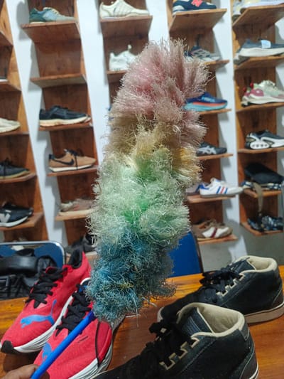 The image captures an indoor scene, likely a shoe store or a personal collection display, filled with various sneakers on wooden shelves against a white wall. In the foreground, a hand holds a vibrant, multi-colored feather duster, transitioning from pink and light brown at the top, through yellow and green, to blue at the bottom. The duster is positioned centrally, partially obscuring the background.

On a wooden table in the immediate foreground, a pair of red Puma running shoes with black laces and blue Puma logos are prominently displayed to the left. Faint text, possibly "ULTIMATE," is visible on the side of one shoe. To the right, a pair of dark, high-top sneakers with light beige interior lining also rest on the table.

The background features numerous pairs of sneakers neatly arranged on wooden shelves of varying heights. Brands such as New Balance, Nike, Adidas, Asics, and others can be identified by their distinctive designs and logos. The shoes exhibit a wide range of colors and styles, from classic white and black to more vibrant combinations. Some appear to be athletic shoes, while others are more casual. The arrangement suggests a curated display. The overall lighting appears to be artificial, illuminating the shelves and shoes. The setting provides a clear view of a diverse collection of footwear.