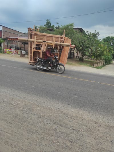 This image, taken in Lancang Barat, Indonesia, captures a man on a motorcycle transporting a large wooden structure along a paved road. The photograph is oriented vertically, with the road running horizontally across the right side of the frame and the sky dominating the left.

The central subject is a man, dressed in a red-brown shirt and dark trousers, seated on a dark-colored motorcycle. Attached to the motorcycle is an elaborate, light-toned wooden framework, extending significantly around the vehicle, suggesting it's designed to carry a substantial load or serve as a mobile stall. Bundles of rolled materials, possibly mats or thin wooden poles, are secured to the top of this wooden structure with red and white ties.

In the background, on the side of the road, are several low-rise buildings, some constructed from red brick, partially visible among abundant green trees and foliage. Overhead, thin power lines stretch across the clear sky, which features scattered white clouds. The asphalt road has a faded yellow dividing line, and the unpaved shoulder consists of dirt and gravel.

A visible banner or sign attached to one of the background buildings displays text advertising food: "MIE CELOR & AYAM GEPREK," indicating the presence of local food vendors. The scene depicts a common form of resourceful transport and commerce in a rural or semi-urban Indonesian setting, occurring during bright daytime hours.
