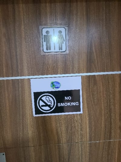 The image displays two laminated or paper signs affixed to a vertical, medium-brown wooden surface, likely a door or wall, in Ibeju, Nigeria. A thin, horizontal white strip runs across the wooden surface, approximately midway down the image. The upper sign, positioned centrally, is a white rectangle with a dark background featuring two white silhouettes: a male figure on the left and a female figure on the right, separated by a vertical line, indicating a unisex or shared restroom. Below this, centered on the surface and partially obscured by the white horizontal strip, is a larger rectangular sign. This lower sign has a black background with white text and symbols, bordered in white. On its left, it prominently displays a "NO SMOKING" symbol—a white cigarette with smoke crossed out by a diagonal line within a circle. To the right of this symbol, the words "NO SMOKING" are printed in white capital letters. A small, circular logo, featuring blue and green colors with what appears to be an island and palm trees, is situated above the black portion of the "NO SMOKING" sign. No individuals are visible.