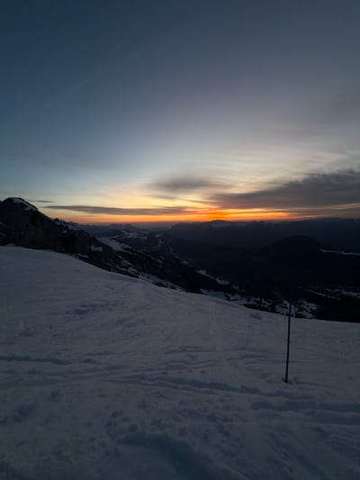 The image shows a sunset over a mountain range, likely taken from a high elevation in La Giettaz, France. A snow-covered slope occupies the foreground. In the middle ground, there is a jagged mountain ridge. The sky is illuminated by a gradient of colors from dark blue to orange and yellow near the horizon. A blue metal pole or ski marker is visible on the right side of the image. The scene is dimly lit.