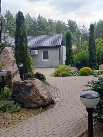A paved pathway winds through a garden with large rocks and manicured bushes. A small, gray building with a dark roof and a single window is visible in the background, along with a tall evergreen tree. Two outdoor lamps are positioned along the pathway, one partially obscured by a large rock. The scene appears to be in a residential area within the Starobin rural council in Belarus.