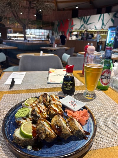 A table is set for a meal at a restaurant, likely in Șelimbăr, Romania, given the context. In the foreground, a blue ceramic plate holds a serving of sushi rolls, garnished with lime wedges, wasabi, and pickled ginger. A small dish with a floral design is also present. Beside the plate are a bottle of soy sauce, a glass of beer, and a bottle of Beck's beer. In the background, the restaurant's interior is visible, with tables, chairs, and people mingling. A tree-like decoration and a mural with bamboo motifs suggest an Asian-inspired theme for the establishment. The lighting appears to be ambient, creating a warm and inviting atmosphere.