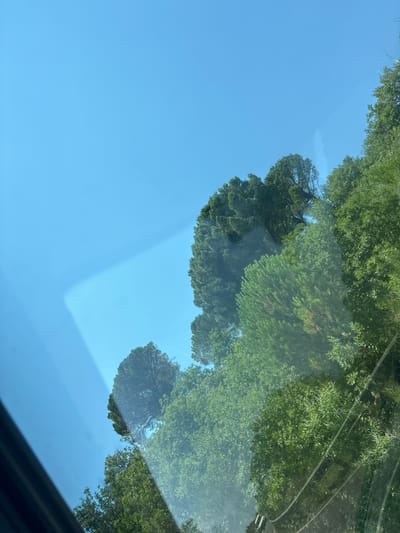 The image shows a clear blue sky with the tops of green trees visible along the right and lower portions. There is a diagonal reflection across the sky, appearing to be from a window. The scene is outdoors, with the sky and foliage indicating a natural environment. The location is identified as Gornji Katun, Montenegro. There are no people or specific actions depicted in the image. The overall impression is a bright, sunny day with a focus on the natural landscape.