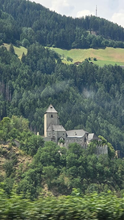The image shows a medieval castle, possibly a fortress, perched on a rocky, tree-covered hillside in Freienfeld - Campo di Trens, Italy. The castle features a tall, square tower with a pointed roof, connected to several other stone buildings with sloped roofs. Battlements are visible along the castle walls. The surrounding environment is dominated by a dense forest of dark green coniferous trees that cover the steep slopes of the mountain. Patches of lighter green grassland are interspersed within the forest. In the far background, the top of a transmission tower is visible on a higher ridge. The foreground of the image is blurred with streaks of green, suggesting movement, possibly from a vehicle. The sky is overcast with some white clouds.