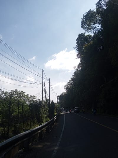 The scene depicts a two-lane asphalt road in Ban Phonhxavanh, Laos, during daylight. A silver metal guardrail runs along the left edge of the road, separating it from an area of dense green vegetation and a series of utility poles supporting numerous overhead power lines. To the right of the road, a steep, heavily forested hillside with dark green foliage casts significant shadow across the roadway. Several vehicles are observed on the road, primarily motorcycles or scooters, with at least one dark-colored four-wheeled vehicle further ahead, all traveling away from the viewer. The sky is bright blue with scattered white cumulus clouds, and the sun, partially obscured by clouds, provides strong backlighting, causing parts of the road and the right-side vegetation to appear in shadow while illuminating the sky and portions of the left-side foliage.