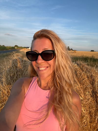 A blonde woman with long, wavy hair and black sunglasses smiles at the camera in a selfie. She is wearing a pink tank top and is positioned in front of a large hay bale. In the background, a rural landscape with a field of wheat, more hay bales, a road, and some houses can be seen under a blue sky with scattered clouds. The lighting suggests it is late afternoon or early evening. The location is identified as Trouville-Alliquerville, France.
