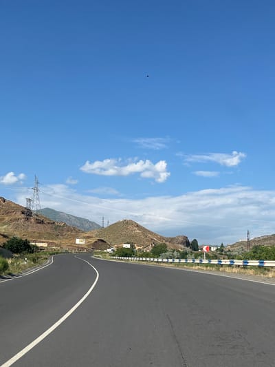 The image shows a scenic view of a winding road passing through a mountainous landscape in Gladzor, Armenia. The road, asphalted and marked with a white line, curves to the right. Along the side of the road, a guardrail with alternating blue and white reflective markers guides the way. The surrounding terrain is characterized by dry, rocky hills with sparse vegetation and some scattered trees.

In the background, a prominent electrical pylon stands tall on a hilltop, with power lines stretching across the frame. Further in the distance, more mountains are visible, some appearing greener than others, suggesting varying altitudes or microclimates. The sky is a clear, bright blue, dotted with a few fluffy white clouds. A small, dark object, possibly a bird, is captured in flight in the upper left portion of the sky.

The lighting suggests it is daytime, likely late morning or early afternoon, with the sun casting shadows that highlight the contours of the land. The overall atmosphere is one of vastness and natural beauty, with a touch of human infrastructure integrated into the environment. There is no visible text in the image.