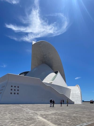 The image depicts the Auditorio de Tenerife Adán Martín in Santa Cruz de Tenerife, Spain. The architectural structure features a prominent curved roof and white concrete construction. A group of four people are walking along the walkway near the building. The sky is blue with scattered white clouds and sun glare. The ground surface consists of grey stone pavement. A piece of construction equipment is visible at the top right of the scene.