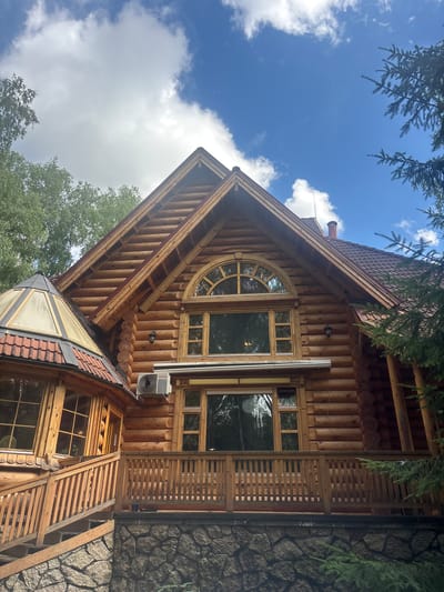 The image shows a multi-story log cabin with a stone foundation, located in Soloslovo, Russia. The building has a large arched window on the upper level and a smaller window below it, both framed by wooden logs. A wooden balcony with a railing runs across the lower part of the main structure. To the left, a separate section of the building features a conical roof with a glass enclosure and a wooden staircase leading up to a window. The exterior also includes an air conditioning unit mounted on the wall, an outdoor light fixture, and a chimney on the roof. Surrounding the cabin are green trees and a blue sky with white clouds.