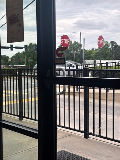 This is an indoor shot from what looks like the inside of a Chick-fil-A restaurant in Stone Mountain, Georgia. Looking out the restaurant door through the metal safety bars, there are stop signs, traffic lights, and street lines painted on the road. The stop signs are partially obscured, with a "Protected by Strong" sign. A Chick-fil-A poster for catering is visible in the left corner. Outside on the street is a Clearinghouse building.