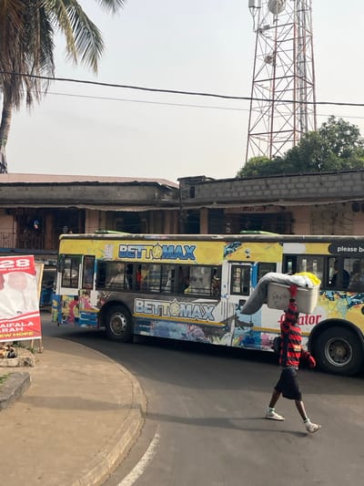 This image depicts an urban street scene in Sumalia Town, Sierra Leone, under bright daylight with a hazy sky. In the foreground, a paved road curves from the left towards the right, bordered by a concrete curb.
A young male individual walks from right to left across the road, positioned in the lower-right quadrant. He wears a red and black horizontally striped long-sleeved shirt, black shorts, white socks, and light-colored sandals. He carries a rectangular, light-gray plastic tub on his head, supported by his raised right arm. The tub contains folded fabrics, including a gray blanket or cloth and other colorful items. Text partially visible on the tub reads "Sumapharm" and "Kekela".
In the midground, a large yellow passenger bus with vibrant, multi-colored graphics and "BETT☆MAX" branding is prominent. The bus appears to be in motion or parked on the road, partially obscuring the background structures. Passengers are visible through its windows.
To the left of the bus, a red and white political campaign poster is visible, displaying a male candidate's portrait and text including "KAIFALA SARAH NEW HOPE" and "MEMBER ASPIRANT," alongside the number "28." A portion of a palm tree is also seen on the far left.
The background features multiple multi-story buildings constructed from concrete blocks, some with corrugated metal roofs, suggesting commercial or residential use. On the far right, a tall, red-painted metal lattice cell tower with multiple antennae extends high into the sky above the buildings. Overhead utility lines are visible crossing the sky. Green foliage is present behind some of the structures.
