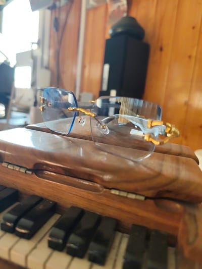 A pair of rimless, blue-tinted sunglasses with gold accents rests on the lid of a wooden object, positioned above piano keys. The sunglasses are the primary subject, with their lenses reflecting the bright, out-of-focus background. The wooden object appears to be part of a musical instrument, possibly an accordion or a small piano, with visible hinges and a textured surface. The piano keys are black and white, partially obscured by the wooden lid. The background is blurry but suggests an indoor setting with wooden paneling on the right and bright lighting from the left, possibly from a window or artificial light sources. There is no indication of the city of Edineț, Moldova, in the image.