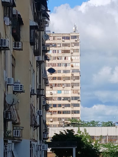 The image depicts a view of apartment buildings in Puerto Ordaz, Venezuela. In the foreground, a pale yellow apartment building with visible air conditioning units mounted on its exterior is partially obscured. Directly behind it, a taller, light-colored concrete high-rise apartment building is prominently featured. This building has numerous windows and appears to be densely populated with air conditioning units and satellite dishes. The sky above is partly cloudy with blue patches, indicating daytime. The scene is urban, characterized by dense residential architecture. No people are clearly visible in the image.