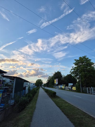 The image depicts a scene in Essenbach, Germany, characterized by a paved sidewalk running alongside a road. The sky is a prominent feature, displaying a blue expanse with scattered white clouds that are illuminated from below by the setting sun, casting warm hues. Three dark power lines traverse the sky diagonally from the upper left to the lower right.
To the left of the sidewalk, a low hedge runs along a grassy embankment. Beyond the hedge, a metallic shed-like structure with a sloped roof is visible. Stacks of colorful containers, possibly for storage or transport, are piled against the side of the structure. A chain-link fence borders this area. Further down the road, more buildings and trees are discernible.
On the right side of the road, there is a line of trees, with one particularly large, leafy tree in the foreground. A street lamp stands near this tree. A paved road stretches into the distance, with a grassy strip separating it from the sidewalk. In the background, several yellow signs with text are posted, and a truck with a crane is parked near one of these signs. The overall impression is one of a suburban or industrial edge of town at dusk.