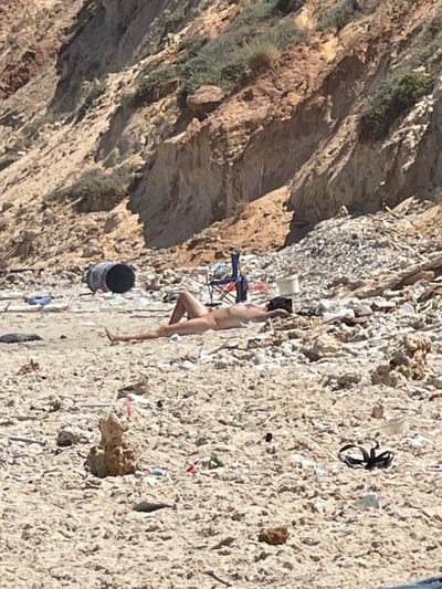 The image depicts a rocky, debris-strewn beach area near the cliffs of Ga'ash, Israel. In the foreground, a person is lying on their back on the sand, with their legs extended. They are unclothed. The beach is littered with various objects, including a large black barrel to the left of the person, a blue cooler, a collapsed camping chair, and piles of what appears to be rubble and trash. The background shows a steep, eroded cliff face with sparse vegetation. The overall scene suggests a neglected and possibly polluted coastal environment.