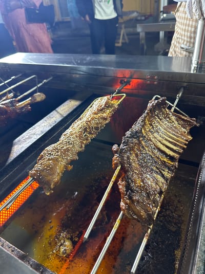 This image shows two skewers of meat being grilled over a hot element in a food stall. The meat on the left skewer is a long, narrow cut, possibly a lamb rib, coated in sesame seeds and a glossy sauce. It is positioned over a red-hot element. The meat on the right skewer is a rack of ribs, also appearing to be lamb, with visible bones and a dark, caramelized crust. It is also coated in sesame seeds and sauce. Both skewers are suspended over a metal tray that is collecting dripping fat and juices, which are bubbling and sizzling from the heat. In the background, blurred figures of people are visible, suggesting this is a public area, likely a street food market. The overall scene suggests a lively food preparation environment.