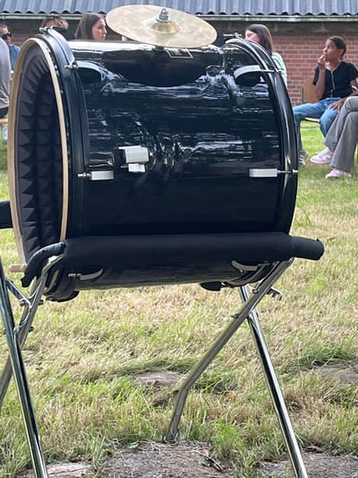 A black bass drum is displayed outdoors in what appears to be a grassy area, possibly a park or backyard. The drum is shiny and black, with metal accents and a cymbal attached to its side. The drum is supported by silver metal stands. The background shows a brick wall and a fence, suggesting an outdoor setting near a building. Several people are visible in the background, some of whom are partially obscured. One person is wearing jeans and pink sneakers, and another is wearing a black t-shirt. The overall scene appears casual, and the lighting suggests it is daytime, possibly late afternoon given the shadows. The grass is dry and sparse in some areas, indicating it might be a warm or dry season. The image is oriented vertically. There is no visible text in the image.