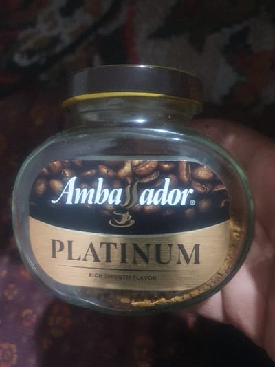 The image displays a glass jar of instant coffee, held horizontally by a person's hand. The jar is filled with light brown, granulated coffee, visible through the clear glass. A prominent label wraps around the jar, featuring a dark background adorned with an image of roasted coffee beans.
Visible text on the label includes the brand name "Ambassador" written in a white, stylized script font on the left side, accompanied by a registered trademark symbol (®) and a small coffee cup icon. To the right of the "Ambassador" logo, the word "PLATINUM" is vertically displayed in large, gold, sans-serif capital letters. Below "PLATINUM," the phrase "RICH SMOOTH FLAVOR" is written in smaller text.
The jar has a dark brown or black lid, with a thin yellow or gold band visible just beneath it. The background of the image is dark and out of focus, showing indistinct patterns that suggest an indoor setting, possibly on a patterned fabric or carpet. Part of a hand, with fingers visible, is holding the jar. No specific time of day or other environmental details are discernible from the image itself. The photograph provides a clear view of the coffee product.