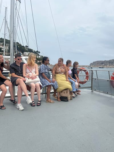 The image depicts seven individuals seated on the grey deck of a passenger vessel, likely a tour boat, within a harbor or coastal waters near Torroella de Montgrí, Spain, specifically consistent with the port of L'Estartit. The sky is overcast.
**Individuals (from left to right):**
1. A partially visible female, dark-haired, wearing a dark top, with her right fist resting on her lap.
2. A male wearing sunglasses, a striped short-sleeved shirt, dark shorts, and white sneakers, seated with his right ankle crossed over his left knee.
3. A female with blond hair, wearing a light-colored, possibly white, patterned strap dress and black sandals, holding a light-colored handbag.
4. A female with dark hair tied back and sunglasses, wearing a blue and white patterned short-sleeved dress and brown sandals.
5. A female with light brown hair, wearing a patterned halter-neck top and a long, pleated yellow skirt. Tattoos are visible on her left arm. She smiles and looks towards the female to her left (Individual 4), engaging in interaction. A tan backpack rests on the deck between her legs.
6. A child, likely female, with brown hair, wearing a light-colored short-sleeved top and patterned blue trousers, facing forward.
7. A child, likely male, with brown hair, wearing a dark short-sleeved shirt, facing forward and looking right.
**Vessel and Environment:**
The individuals are seated on a metal bench structure. The boat's port side reveals multiple ropes (rigging) and a mast. The starboard side features a metal railing with clear panels and two orange and white lifebuoys. The deck surface is a uniform grey. In the background, to the port, a vegetated hillside is covered with numerous white buildings of a coastal town. To the starboard, calm blue water shows several smaller boats anchored. A rocky headland or island is visible in the distance under the light gray, overcast sky.