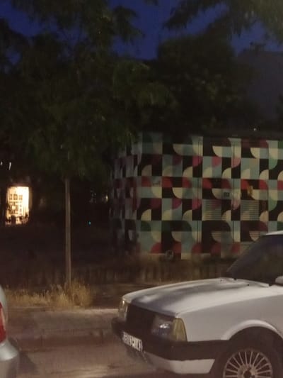The image depicts a nighttime scene in Karşıyaka, Turkey. A white car is prominently featured in the foreground, partially obscuring a sidewalk and a street. Behind the car, a building with a distinctive geometric mural is visible. The mural is composed of squares and semicircles in various colors, including black, white, light blue, and shades of red and pink. To the left of the building, a brightly lit window suggests an indoor space.
The setting appears to be urban, with trees lining the area and dry vegetation along the roadside. The sky above is dark, indicating nighttime. The overall mood of the image is quiet and urban.
There are no clearly discernible people in the image, although faint shapes that could be figures are present within the building behind the mural. The activity taking place is not evident, as the scene is static.
Visible text includes a license plate on the white car, but the characters are too blurry to be read. Another faint text appears on the mural itself, but it is illegible. The time of day is clearly night, as evidenced by the dark sky and artificial lighting. There are no direct cues about the weather, but the general atmosphere suggests a clear night.