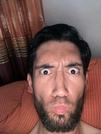 A close-up, selfie-style photograph shows a person's face with a distorted, surprised expression. The person has dark hair, a beard, and prominent eyebrows. Their eyes are wide open, and their mouth is pursed. The background features an orange-striped pillow and a section of rust-colored curtains with gold accents. The lighting suggests an indoor setting, possibly a bedroom. There is no indication of the location being Mashrab, Uzbekistan; this detail cannot be confirmed from the image.