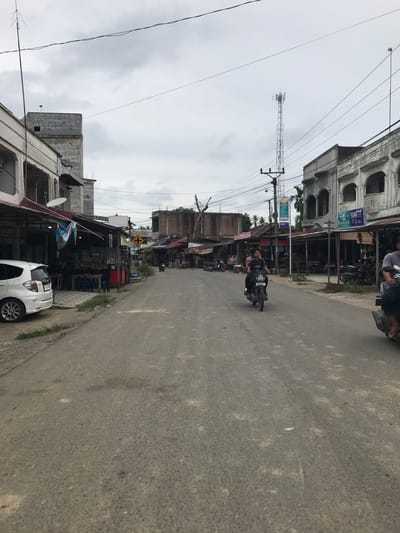 The image depicts a street scene in Keudesimpangempat, Indonesia, oriented with the road running lengthwise. A two-lane asphalt road occupies the majority of the frame. A motorcycle with two riders is in the center of the road, moving toward the camera. The road is flanked by buildings on both sides, mostly one and two-story structures, some with storefronts. The buildings suggest a mix of residential and commercial use. A white car is parked on the right side of the road. Overhead power lines are visible, crisscrossing the sky. A tall communication tower rises in the distance. The sky is overcast. The overall impression is that of a small town.