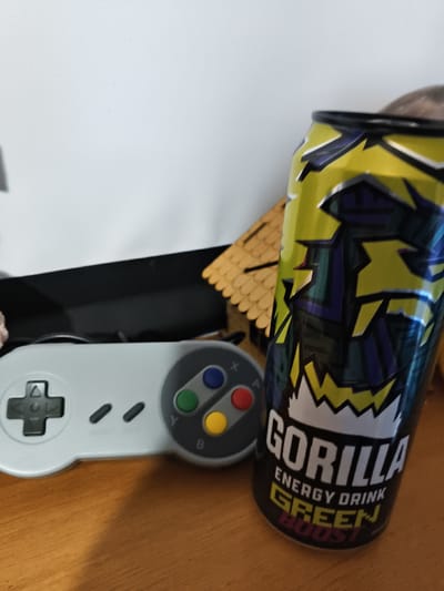 The image shows a close-up of a gaming setup with an energy drink can. In the foreground, a light gray video game controller, resembling a Super Nintendo controller, is positioned on a wooden surface. The controller has a directional pad on the left and four colored buttons (green, blue, red, yellow) on the right, labeled Y, X, B, and A respectively. Two small oval indentations are visible between the directional pad and the buttons.

Next to the controller, to its right, is a tall, slender can of "GORILLA ENERGY DRINK GREEN BOOST." The can has a predominantly yellow and green design with sharp, angular graphics in purple and black, evoking a sense of energy and intensity. The text "GORILLA," "ENERGY DRINK," "GREEN," and "BOOST" is clearly visible on the can.

In the background, a small, stylized wooden house-like structure with a brown, shingled roof is partially visible. Behind the objects, a plain white wall forms the backdrop. The lighting suggests an indoor setting, possibly during daytime, with soft illumination. There are no people visible in the image. The scene evokes a casual atmosphere associated with gaming and energy consumption. The location is identified as Mokshan, Russia.