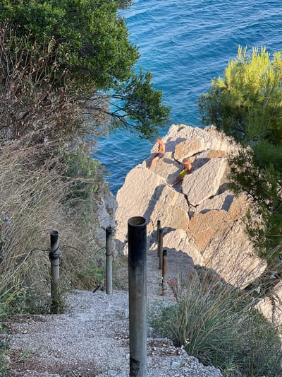 The image shows a rocky coastline in Medinski Krš, Montenegro. Turquoise water can be seen below a rocky cliff. Two people lie prone on the cliff's edge. A series of metal pipes are attached to a steep path leading away from the cliff. Vegetation including trees, shrubs, and grasses is visible. The scene appears to be taken from a high vantage point.