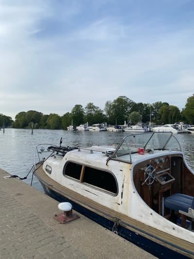 The image shows a close-up of a docked boat in Elmbridge, United Kingdom. The boat is primarily white with a wooden interior and a blue trim along the edge. It appears to be an older model and is tied to a gray concrete dock. In the background, a line of similar boats is visible, docked along the tree-lined riverbank. The sky is overcast. A metal mooring bollard is visible on the dock next to the boat.