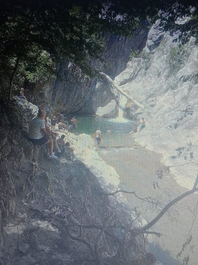 The image depicts a natural scene of a waterfall cascading into a pool, surrounded by rocky terrain and lush vegetation. Several individuals are visible, some seated on the rocks near the pool and others in the water. A fallen log spans across a portion of the scene, near the waterfall. The environment appears to be a secluded, natural area, possibly a popular spot for recreation. The location is identified as being in the city of Gorno Aleksandrovo, Bulgaria.
