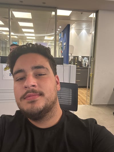 The image depicts a male individual in an office environment, described as being located in Azor, Israel. The subject, a man with dark hair, a mustache, and stubble, is shown from the chest up, wearing a plain black t-shirt, looking directly at the camera with a neutral expression. He is seated, with a black mesh-backed office chair visible behind him to the right.
The background shows a modern office setting. To the subject's left, a white office cubicle partition with a yellow-and-black bee logo featuring the number "8" is visible. Reflections in clear glass partitions above and behind the cubicle reveal another individual wearing glasses, partially visible, and additional open-plan office spaces with overhead fluorescent lighting.
To the subject's right, a partially open clear glass door leads into an adjacent room. This room appears to be a kitchenette or break area, featuring dark gray base cabinets with horizontal pull handles, a light-colored countertop, a golden-toned faucet, a white wall-mounted paper towel dispenser, and several white and clear beverage machines or dispensers. The floor in this area is a light-colored wood laminate. A blue partition or wall with white text is also visible within this room.