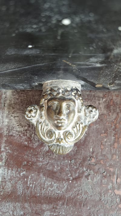 A close-up, vertical shot shows a decorative brass face adorning the side of an old, possibly wooden, dark-colored surface, which appears to be attached to a weathered, reddish-brown wall. The brass face is that of a woman with a laurel wreath on her head. Her eyes are closed, and her mouth is slightly parted. On either side of her head are floral ornaments. The brass has a patina, indicating age. The surface above the face is dark and appears to be a metal overhang or lintel with some diffused lighting, possibly indicating an outdoor setting. The wall below is rough and shows signs of wear and possibly paint chipping. The image does not provide any specific details that would allow for precise location identification within the city of Fontainebleau, France.