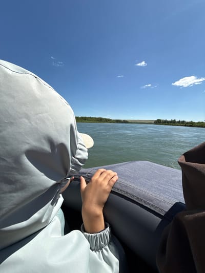 In Belyi Yar, Russia, a child's hand rests on a blue inflatable raft on a river. The child is wearing a light blue hooded garment, partially obscuring a beige baseball cap. The raft is positioned on the water, with the child facing away from the camera. The river is wide and calm, with a tree-lined shore visible in the distance under a clear blue sky with scattered white clouds. The perspective is from within the raft, looking out at the landscape.
