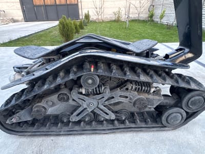 A black, tracked vehicle attachment, likely for a snowmobile or all-terrain vehicle, is centrally positioned on a light gray concrete surface. The track system is substantial, revealing numerous suspension components including springs, multiple rollers, and an X-shaped structural bracket. Two black footrests, textured with small studs, are mounted on the upper frame of the track unit. A distinct, red-tipped metallic object, resembling a bullet casing or a small bullet, rests on one of the track treads.
The background features a well-maintained grassy area with several small evergreen shrubs and a few bare deciduous trees, suggesting early spring or late autumn. Behind the greenery, there is a light-colored concrete block wall or fence, and in the distance, a large, dark gate, possibly wooden or metallic. The setting appears to be a backyard or private outdoor space in Misaktsieli, Georgia.
The image is taken during the daytime, under clear weather conditions, indicated by the soft, even natural light. No people are present, and the vehicle is stationary, indicating no current activity. A small blue sticker with unreadable white text is partially visible on the side of the track's frame.