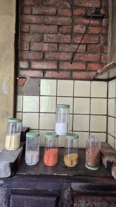 This is an image of a rustic kitchen area, likely in Mashezero, Russia. The scene features a brick wall on the left, some of which is covered in a light-colored tile with a diamond pattern. Several clear plastic jars with green lids are arranged on a flat surface on the right. These jars contain various dry goods: the top jar holds what appears to be buckwheat or a similar grain, the second contains yellow lentils or split peas, the third has orange lentils, and the fourth contains a white granular substance, possibly sugar or salt. The bottom two jars are partially visible and appear to contain more dry goods, one yellowish and the other white.
Next to the tiled area, on the right side of the image, is a dark metal structure that resembles an old stove or oven, with signs of rust and wear. A metal rod is attached to the brick wall, extending outwards. The overall impression is one of an older, functional kitchen, possibly in a rural or traditional setting. The lighting suggests it's daytime, but there are no strong indications of weather conditions. The image is oriented vertically. No people are visible. There is no visible text in the image.