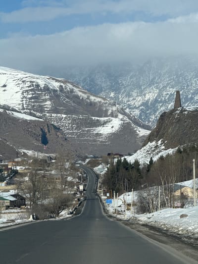 This image depicts a snow-covered mountainous landscape in Sioni, Georgia, viewed along a two-lane asphalt road receding into the distance. The road, appearing damp or recently cleared, curves gently to the left and undulates over a rise. On the right, a prominent stone defensive tower stands atop a steep, rocky, snow-dusted outcrop. The surrounding mountain slopes are rugged, with significant patches of snow contrasting with exposed dark rock and sparse vegetation.

To the left of the road, a collection of single and two-story buildings, characteristic of a village or small town, are visible, partially obscured by bare trees and light snow cover. A Georgian flag, red and white, flies from a flagpole on the left side, slightly elevated above the road. More buildings are sparsely distributed on the right side of the road, alongside bare deciduous trees and some conifers. Utility poles and some commercial signage, including one mentioning "HOTEL CAMPING," line the road on the right.

The sky above the distant mountain peaks is partially cloudy, with a lighter blue showing through in the upper portion. No individuals are visible in the scene. A dark vehicle is discernible in the distance on the road, heading away from the viewer. The overall environment suggests cold, possibly winter or early spring conditions.