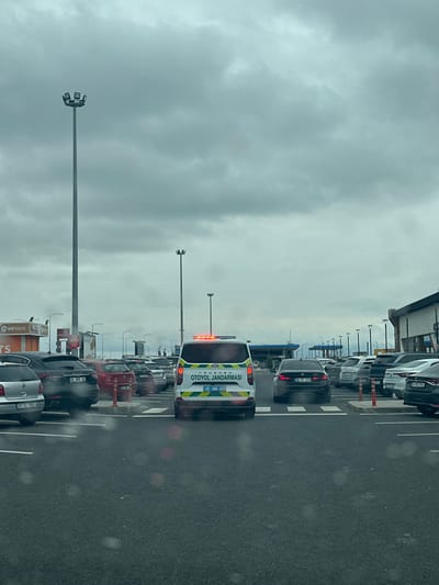 The image depicts an outdoor parking area in Kemalpaşa, Turkey, under an overcast sky. A white Ford Transit-style van, marked "OTOYOL JANDARMASI" (Highway Gendarmerie) with blue lettering and a green and light blue stripe, is centrally positioned, facing away from the viewer. The gendarmerie vehicle has illuminated rear lights and a red flashing light bar on its roof, displaying the license plate "45 JAA 422". It is situated on a white-striped pedestrian crossing. Numerous private passenger vehicles, including sedans, SUVs, and hatchbacks, are parked in marked spaces on both sides of the gendarmerie van, extending into the background. Visible license plates include "35 UEB 35", "35 TC 888", "34 BT 319", and "35 PCJ 942". The ground consists of dark asphalt pavement with white painted lines for parking and pedestrian crossings. Several tall, grey light poles with multi-fixture lamps are present throughout the scene. In the background, structures indicative of a service area are visible, including a blue canopy likely belonging to a fuel station, and a partial sign for "DTS Türkiye" on the left. No individuals are clearly visible.